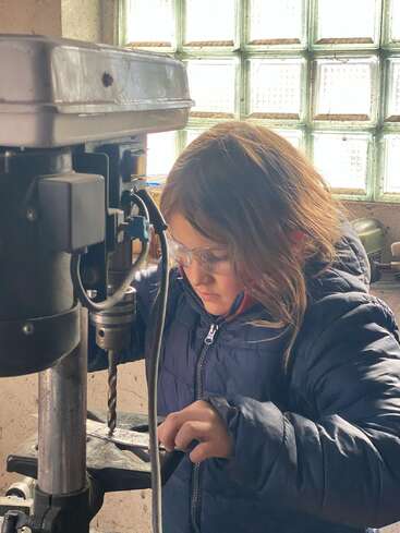 Une jeune fille portant des lunettes de sécurité et une veste bleue utilise une perceuse à colonne dans un atelier, se concentrant sur sa tâche, entourée d'équipements industriels.