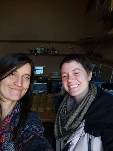 Two smiling women take a selfie indoors, standing in a cozy kitchen with shelves, jars, and kitchen items in the background, creating a warm, friendly atmosphere.