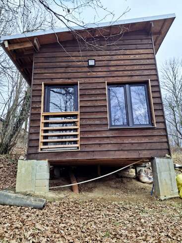 This image shows a small wooden cabin elevated on concrete blocks, surrounded by fallen autumn leaves and bare trees, with two large windows and a simple sloped roof.