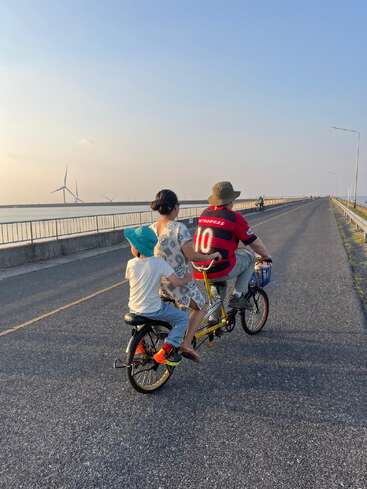 Three people ride a tandem bicycle on a sunny road by the seaside. Wind turbines stand in the background, capturing the peaceful outdoor moment together.