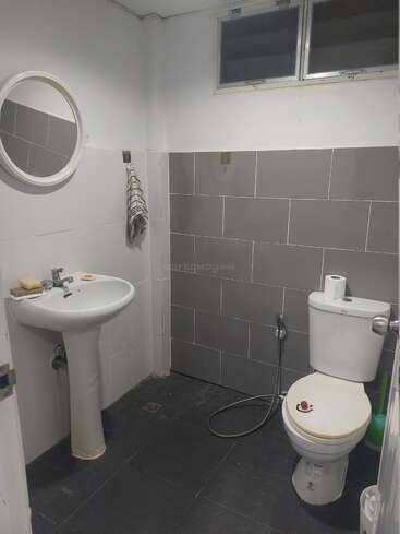 This bathroom features a pedestal sink, round mirror, towel, toilet with a bidet spray, toilet paper, cleaning brush, and grey and white tiled walls.