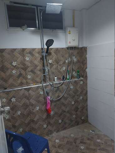 This image shows a bathroom with brown patterned tiles, a blue plastic chair, a wall-mounted shower, water heater, window, toiletries, and a pink loofah hanging.