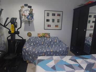 This cozy room features a bed, exercise machine, blue-patterned couch, dark wardrobe with a mirror, wall art, and a small shelf with photos.