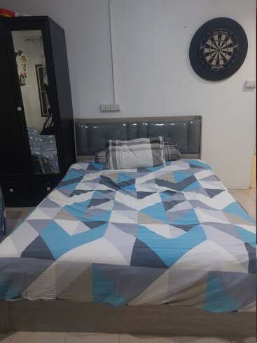 Un lit recouvert d'une couverture à motifs géométriques bleus, blancs et gris est adossé à un mur. Au-dessus se trouve un jeu de fléchettes. Une armoire avec un miroir se trouve à gauche.