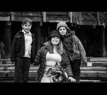 Una fotografía en blanco y negro muestra a una mujer sentada junto a un perro, flanqueada por dos niños pequeños. Todos sonríen, de pie ante unas vigas de madera de estilo rústico.