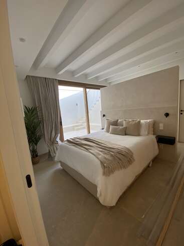 This serene bedroom features a cozy bed with neutral bedding, several pillows, a soft throw, large window, potted plant, beige walls, and minimalist decor.