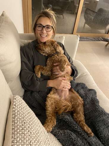 A woman wearing glasses smiles while relaxing on a couch, holding a fluffy brown dog in her lap. They both look cozy with a dark blanket.