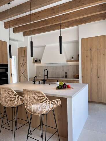 This modern kitchen features wood beams, rattan barstools, a large island with black fixtures, pendant lights, wood cabinetry, and fresh vegetables, creating a warm, inviting space.