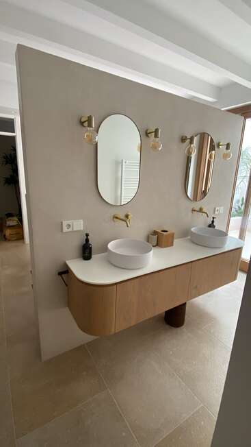 This image shows a modern double sink bathroom vanity with round vessel sinks, wooden cabinets, oval mirrors, brass fixtures, minimalistic decor, and neutral tones, exuding elegance.