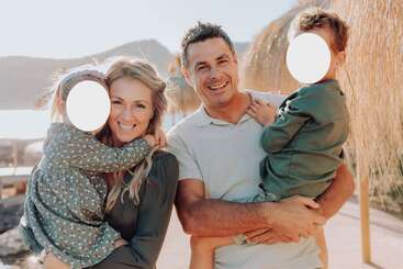 A happy family of four poses outdoors. Parents hold their two young children, smiling in the sunlight with mountains and water in the background—faces blurred.