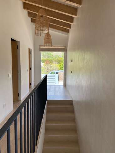 Ein heller Flur mit Holzbalken an der Decke, zwei Hängelampen aus Korbgeflecht, einer beigen Treppe, einem schwarzen Geländer, weißen Wänden und Türen, die zu den Zimmern führen. Hier fällt Tageslicht herein.
