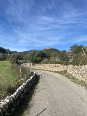 Eine kurvenreiche Landstraße, gesäumt von Steinmauern und grünen Feldern, führt unter einem strahlend blauen Himmel mit vereinzelten Wolken auf die fernen Hügel zu.
