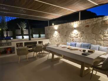 Dieser gemütliche Essbereich im Freien besticht durch eine Steinmauer, eine Holzdecke, einen modernen Grill, gepolsterte Sitzgelegenheiten, eine elegante Tischdekoration und stimmungsvolle Beleuchtung – ideal für abendliche Zusammenkünfte und zum Entspannen.