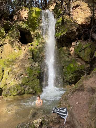 Eine Frau sitzt in einem kleinen Teich am Fuße eines malerischen Wasserfalls, umgeben von moosbewachsenen Felsen, Bäumen und Sonnenlicht; daneben liegt ein gestreiftes Handtuch.