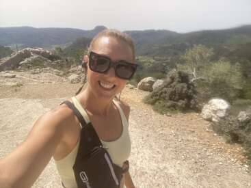 Eine Frau mit Sonnenbrille lächelt für ein Selfie, während sie in der Natur wandert. Sie trägt ein helles Tanktop und eine Umhängetasche und steht inmitten einer malerischen Berglandschaft.