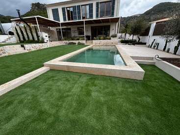 Ein modernes Haus mit großen Fenstern, einer überdachten Terrasse und einem eleganten, rechteckigen Pool, umgeben von Kunstrasen und gestaltet mit Pflanzen und Steinmauern.