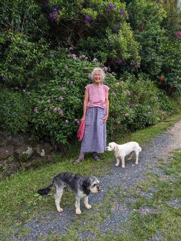 Eine ältere Frau steht lächelnd auf einem Gartenweg, umgeben von üppigem Grün und lilafarbenen Blumen, begleitet von zwei kleinen Hunden, die die freie Natur genießen.