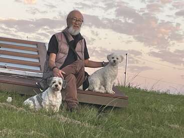Ein älterer Mann mit grauem Bart sitzt bei Sonnenuntergang auf einer Bank im Freien, begleitet von zwei kleinen weißen Hunden, umgeben von grünem Gras und weichen Wolken.