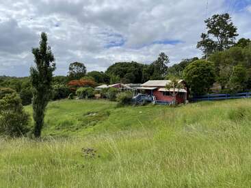 Ein rotes Haus steht auf einem grasbewachsenen Hügel, umgeben von üppigen grünen Bäumen und einem blauen Zaun, unter einem teilweise bewölkten Himmel in einer friedlichen ländlichen Umgebung.