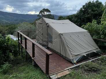 Ein großes Zelt aus Segeltuch steht auf einer hölzernen Plattform, umgeben von üppigem Grün und mit Blick auf die fernen Berge unter einem wolkenverhangenen Himmel, der einen friedlichen, malerischen Platz zum Zelten schafft.