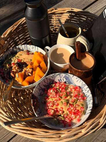 A wicker basket holds two bowls of oatmeal topped with fruit, nuts, seeds; accompanied by tea, coffee, a black thermos, mugs, and sunlight.
