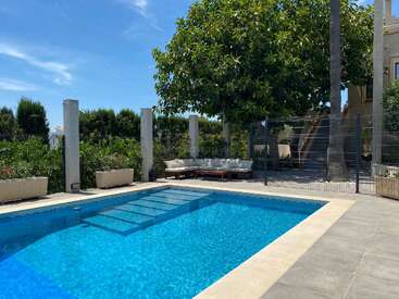 A sparkling blue swimming pool sits beside a relaxing outdoor lounge area under a leafy tree, surrounded by greenery and a sunny, clear blue sky.