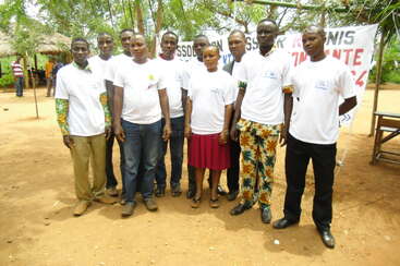 A group of nine people stands outdoors on sandy ground, wearing matching white t-shirts. Trees and a banner are visible in the background, suggesting a community event.