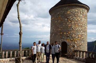 Five people pose together on a stone terrace, standing in front of a circular, stone tower with a domed roof, overlooking a scenic, mountainous landscape.