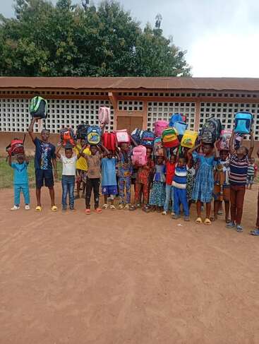 A group of cheerful children stand in a row outside, joyfully holding colorful backpacks above their heads, with a school building and trees in the background.