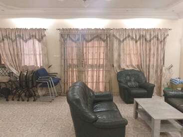 This living room features green leather sofas, a gray coffee table, patterned curtains, stacked chairs, tiled flooring, and cream-colored walls, creating a cozy atmosphere.