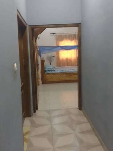 This image shows a hallway leading to a bedroom with a blue mosquito net, a wooden bed, a standing fan, and light curtains covering the window.