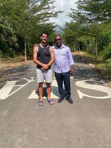 Two men stand together on a road surrounded by trees. One wears casual sportswear and slippers; the other is dressed formally. They both smile, enjoying sunlight.