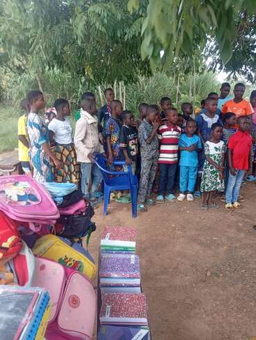 A group of children stands outdoors under a tree. In the foreground, colorful school bags and notebooks are neatly arranged on the ground, suggesting a donation event.