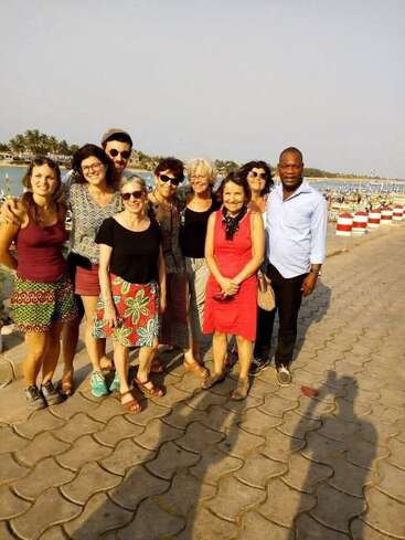 A group of nine people stand together smiling on a waterfront promenade. The sunny weather creates long shadows. Palm trees and water are visible in the background.
