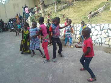 A group of children joyfully dance together outdoors on a concrete surface, watched by others sitting nearby. They appear to be enjoying a festive, lively moment.