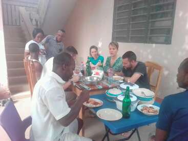 A group of people sits around a table outdoors, sharing a meal. Plates, bottles, and food are on the table. Some people are standing, chatting, smiling.
