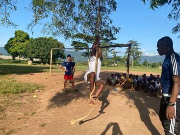 A group of people watch as a person in white climbs a rope hanging from a tree on a sunny outdoor field, surrounded by greenery and mountains.