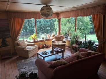 Un salon confortable avec de grandes fenêtres, des plantes et des sièges confortables. La lumière du soleil pénètre à travers les rideaux orange. Un chat est assis paisiblement sur le rebord de la fenêtre.