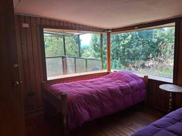 Cette chambre en bois confortable comprend deux lits simples avec des couettes violettes, de grandes fenêtres d'angle offrant une vue pittoresque sur la forêt et une petite table d'appoint ronde en bois.
