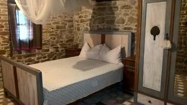 Esse quarto tem paredes de pedra rústica, uma cama de casal com roupa de cama branca, duas mesinhas de cabeceira, um guarda-roupa, uma pequena janela com cortinas escuras e um mosquiteiro pendurado.