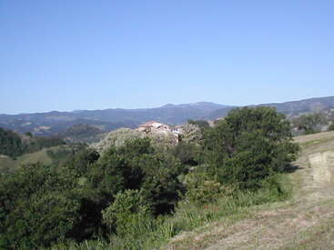 Esta imagem mostra uma paisagem rural tranquila com árvores verdes exuberantes, colinas onduladas e uma casa parcialmente escondida. Montanhas e um céu azul claro completam a cena.