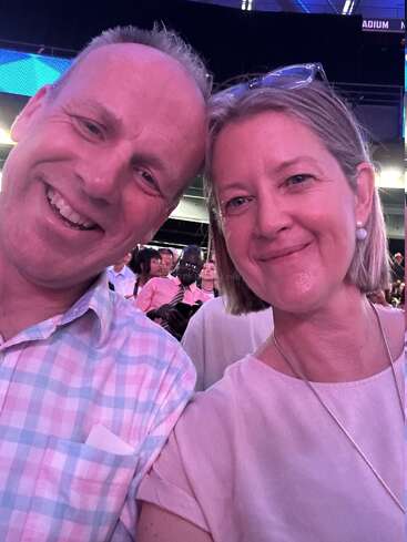 The image shows a selfie of a man and woman posing together in a crowded room, with the woman wearing a pink shirt and the man in a plaid shirt.