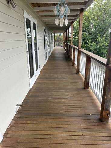 The image depicts a long wooden porch with a railing, featuring white siding on the left and a blue chandelier hanging from the ceiling, surrounded by trees.
