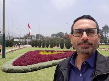 Das Bild zeigt einen Mann vor einem gepflegten Garten mit einem leuchtend roten Blumenbeet und einer peruanischen Flagge, die an einem bewölkten Tag im Hintergrund weht.