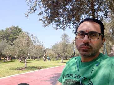 Das Bild zeigt einen Mann in einem grünen T-Shirt, der an einem sonnigen Tag vor einem Park steht, mit Bäumen, Gras und einem gepflasterten Weg im Hintergrund.