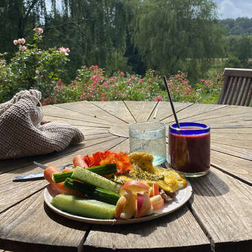 En una mesa de madera al aire libre hay un plato con verduras frescas, una tortilla y rodajas de manzana, junto a unas bebidas. Al fondo, un jardín soleado con flores en flor.