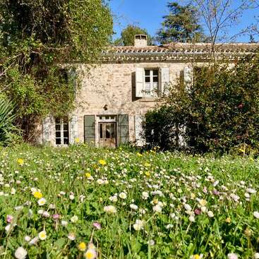 Uma charmosa casa de pedra com venezianas verdes fica atrás de um jardim exuberante, repleto de flores silvestres e grama alta, cercada por árvores sob um céu azul claro.
