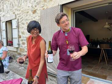 Two people are standing outside by a stone wall, smiling and holding glasses of red wine. One person also holds a wine bottle, enjoying a casual gathering.