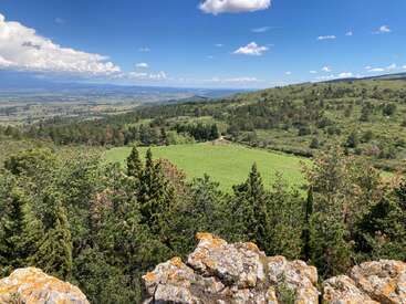 A breathtaking landscape featuring rocky outcrops in the foreground, lush green trees, rolling hills, a sunlit meadow, and blue sky with scattered clouds above.