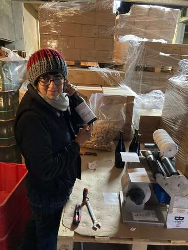 A person wearing a red and gray knit hat, glasses, and scarf labels wine bottles in a warehouse surrounded by stacked boxes, corks, and bottling equipment.
